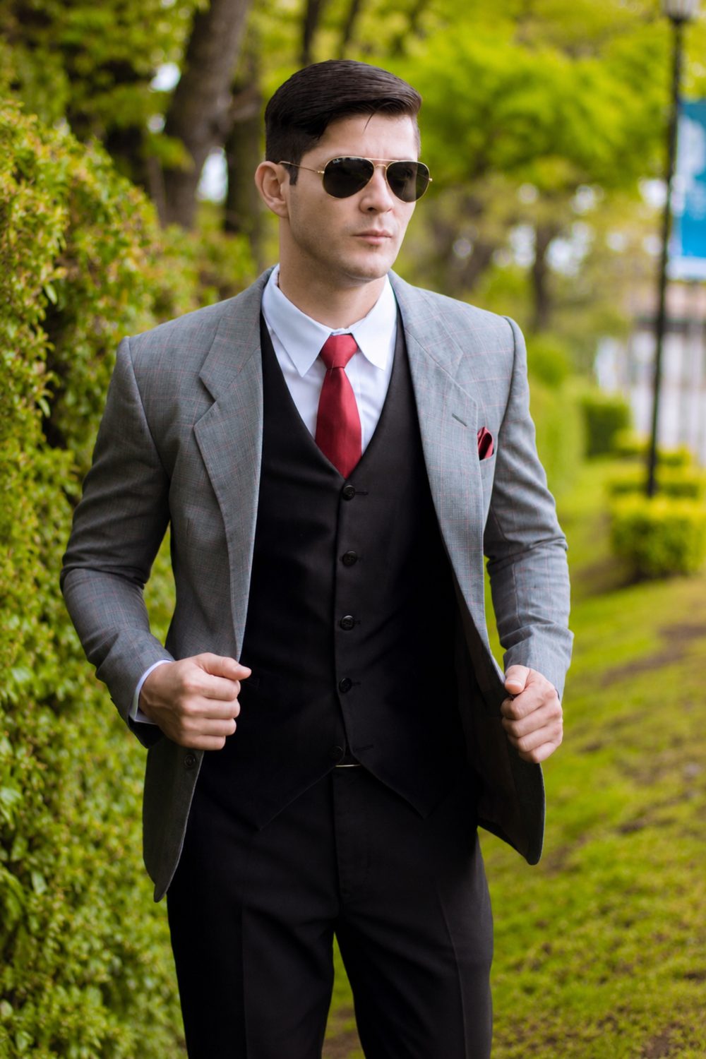 Caballero con saco gris cuadriculado, chaleco negro y corbata roja, lentes de sol en parque