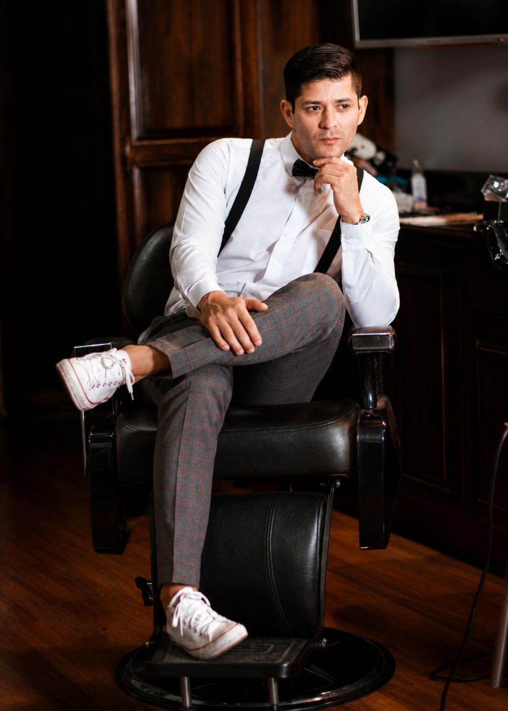 Caballero con pantalón a cuadros grises, camisa blanca, tirantes y corbatín negro en silla de barbería