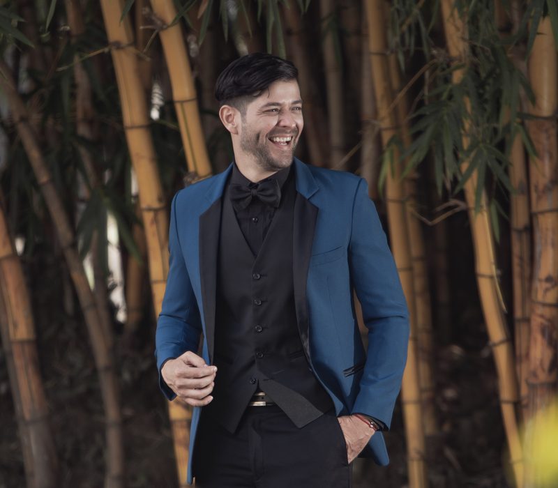Caballero sonriendo con smoking azul petróleo y corbatín negro entre bambúes