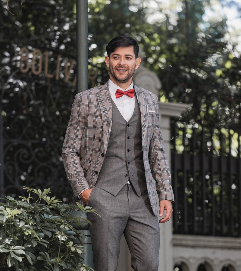 Caballero con saco escocés beige, chaleco gris y corbatín rojo en jardín